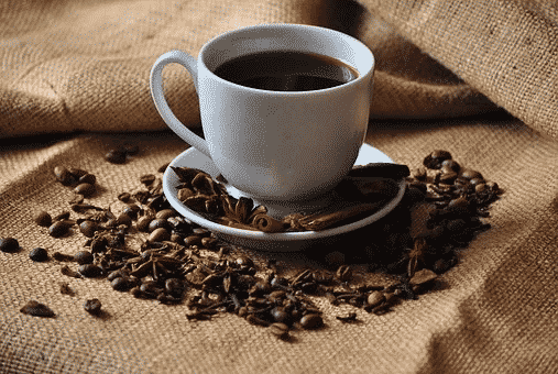 A white cup full of black coffee with a sack in the background and spices and coffee beans scattered around it