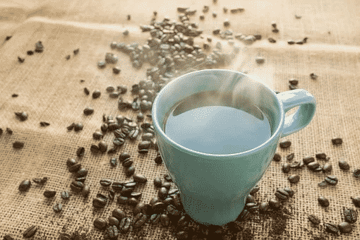 A blue mug full of black coffee with steam rising from it and coffee beans in the background
