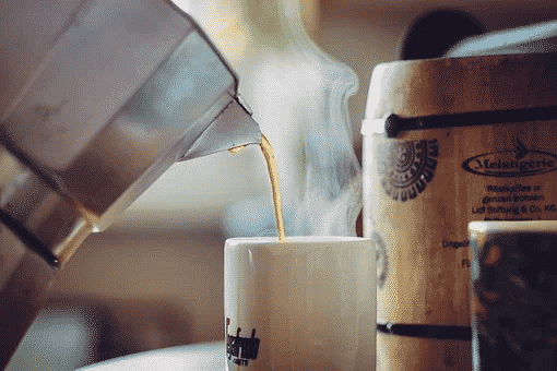 A kettle pouring hot coffee in a white mug with steam rising on top