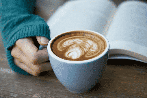 A person in a blue sweater holding a white cup of coffee infron of an open book